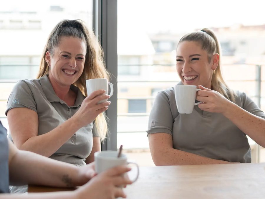 Zwei Kolleginnen trinken lachend Kaffee am Fenster